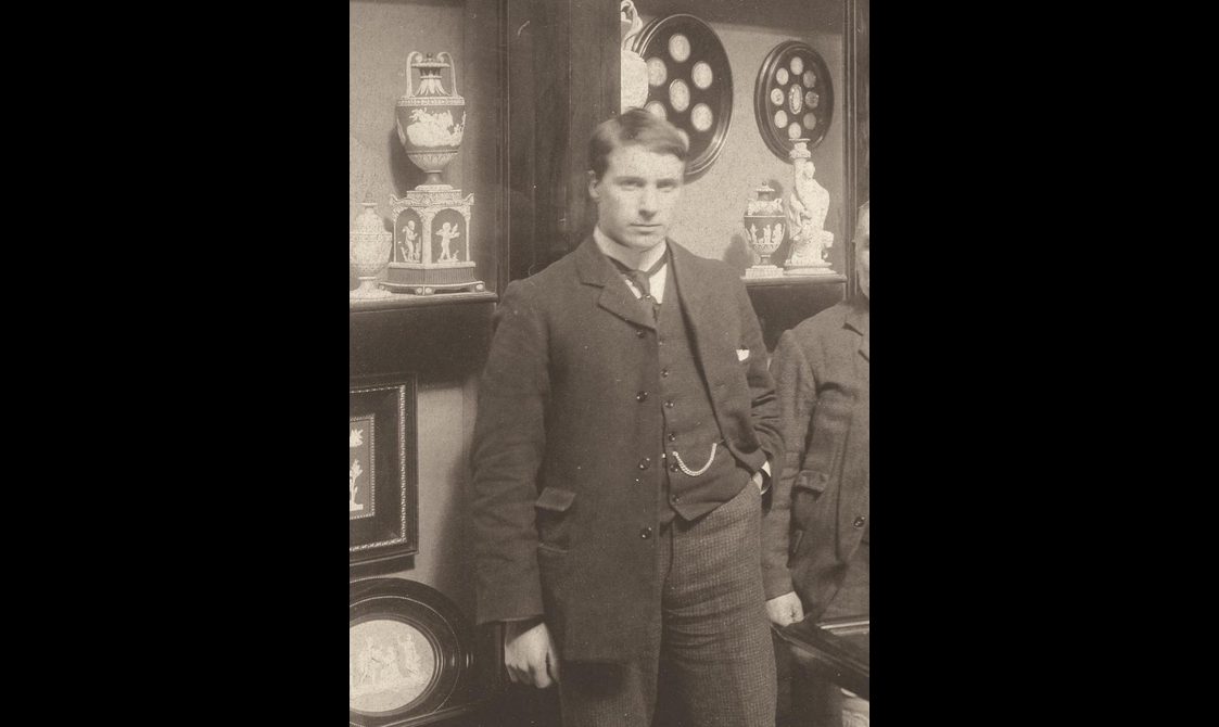 Sepia toned photograph of a man standing in a three-piece suit in front of some shelves of objects.