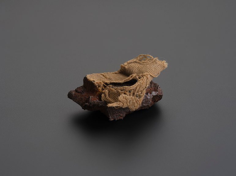 Fragment of metal with some woven fabric attached to it.