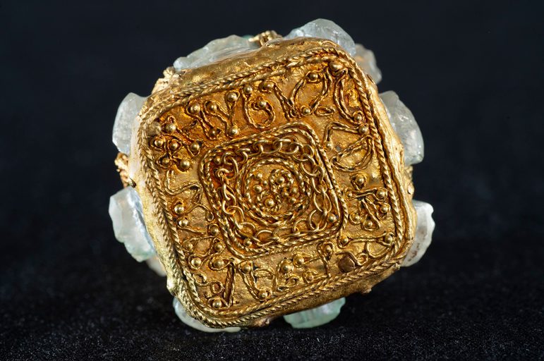 The golden base of a jar, turned to face forward. Golden lines form letters going around the base's square edges.