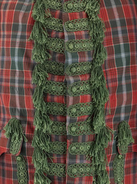 A close up of a red and green tartan jacket. Elaborate green embroidery and tufts  are placed around the green buttons in the center of the jacket.